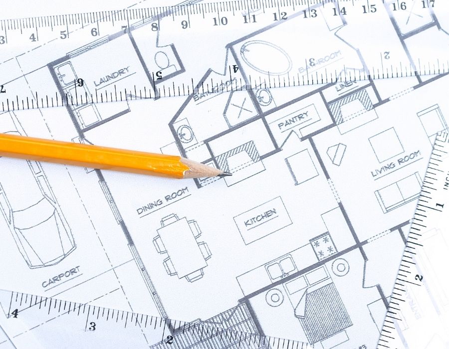 Custom home floor plan with labeled kitchen, dining room, living room, and carport, shown with architectural scale rulers and a yellow drafting pencil.