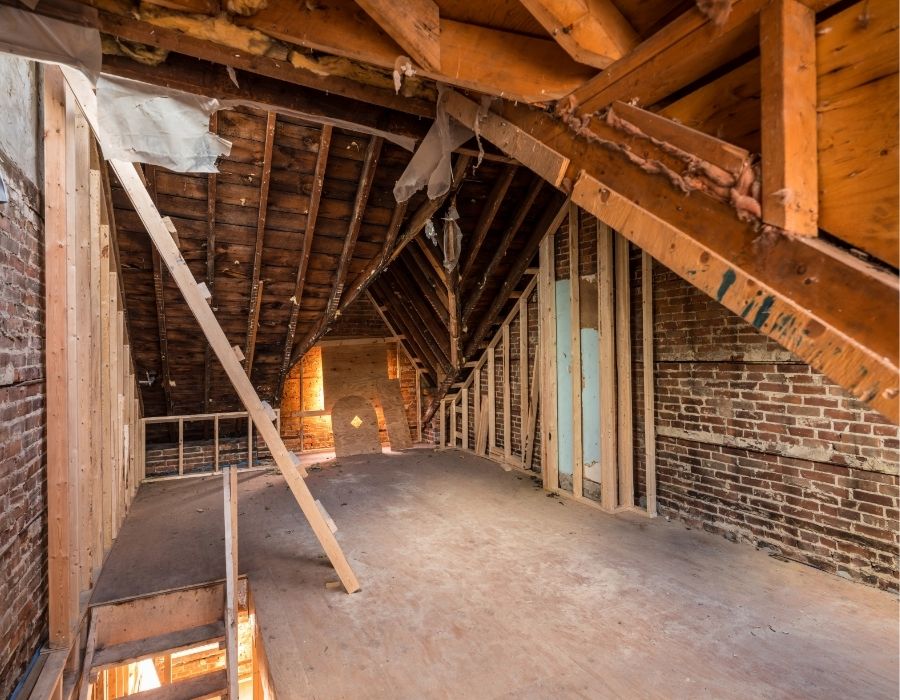 Unfinished attic renovation with exposed wood framing, brick walls, and pitched ceiling in an old house undergoing structural restoration.
