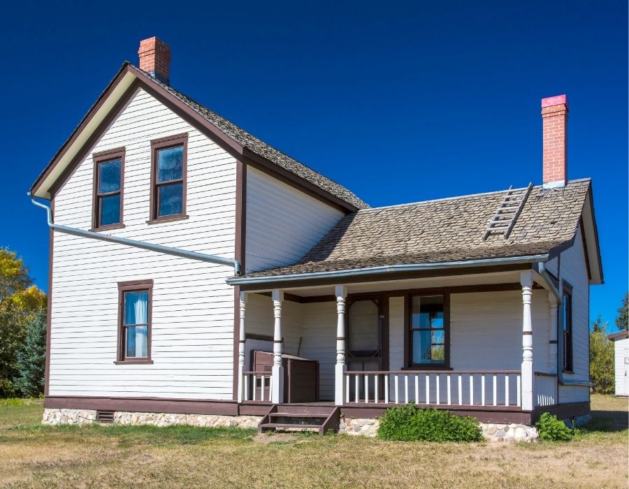 Historic two-story farmhouse with wood siding, front porch, brick chimney, and shake roof, showcasing vintage rural architecture under a clear blue sky.