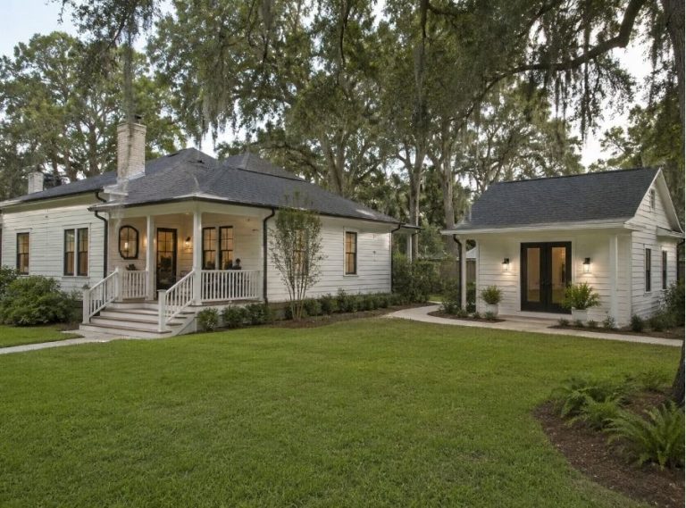 Modern Savannah, Georgia backyard featuring a detached ADU designed to match the main Southern-style home with white siding, dark roof shingles, covered porches, landscaped lawn, mature oak trees, and Spanish moss creating a cohesive residential property aesthetic.